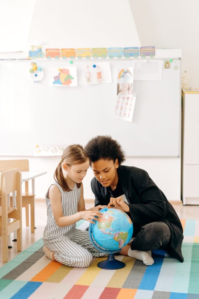 A teacher and a child study a globe together in a bright classroom setting.